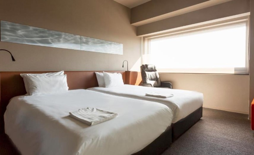 Twin room with horizontal wave-pattern wall panel, leather headboard and full-length window. Reclining chair in corner