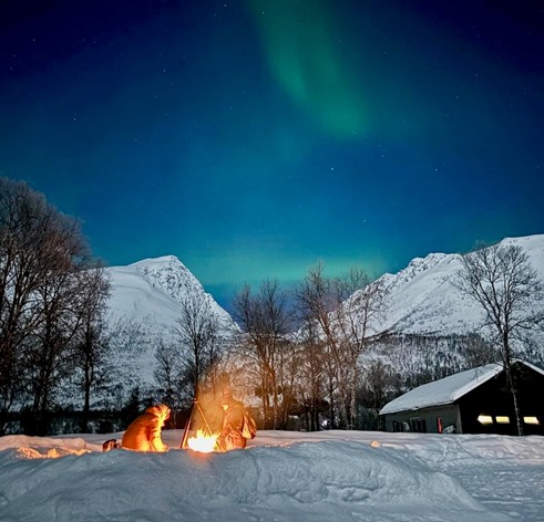 Magical winter night scene in Tromsø, featuring a cozy campfire in the snowy foreground, with the Northern Lights dancing across the sky above snow-capped mountains and bare trees.