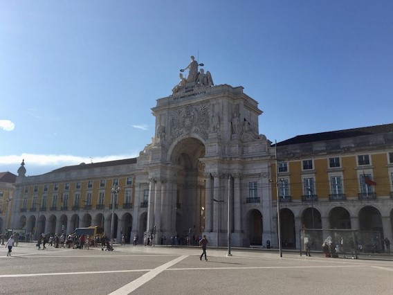 Praça do Comércio in Lisbon, featuring the grand Arco da Rua Augusta triumphal arch and yellow Pombaline-style buildings. People walk across the large plaza under a clear blue sky.