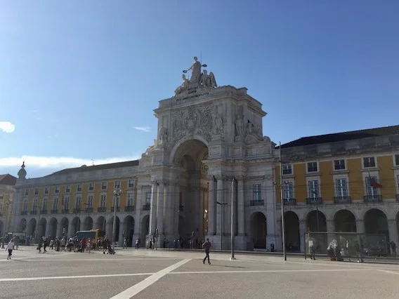 Praça do Comércio in Lisbon, featuring the grand Arco da Rua Augusta triumphal arch and yellow Pombaline-style buildings. People walk across the large plaza under a clear blue sky.