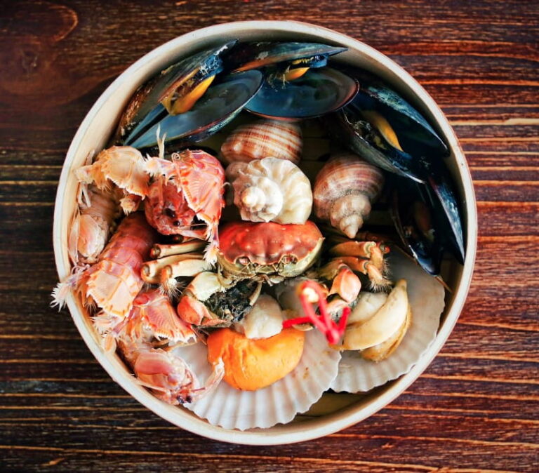A vibrant and colorful assortment of seafood in a bowl, featuring mussels with dark blue shells, several spiral-shelled snails, orange crab pieces, and what appears to be a small lobster or crayfish. The bowl is placed on a wooden surface with rich brown tones that complement the warm hues of the seafood. The image captures the variety and freshness of the seafood selection, making it visually appealing and suggesting a gourmet dining experience