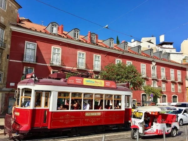 A traditional tram car traveling through the historic Baixa district of Lisbon, passing by the colorful buildings and lively street scene. With a tuk tuk in the foreground