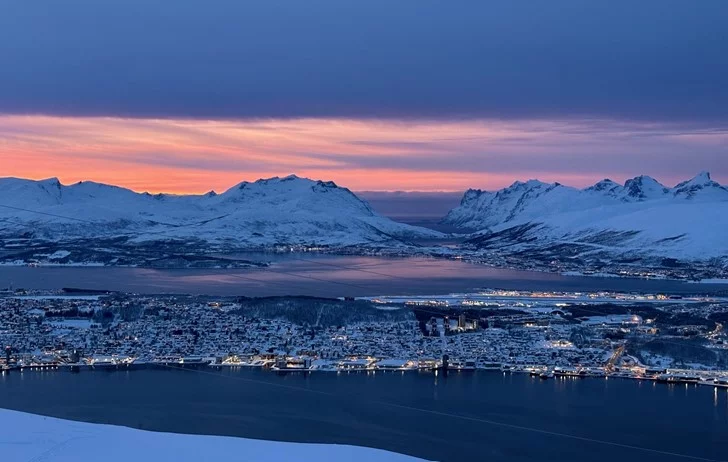 Breathtaking dusk view of Tromsø from above, with city lights twinkling below snow-covered mountains and dramatic orange-pink sky.