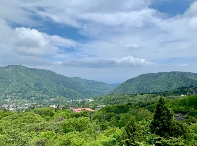 Scenic view of the lush green mountains and valley of Hakone, with a town nestled among the trees and partly cloudy blue sky overhead