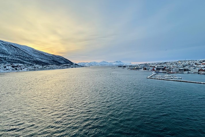 Scenic fjord view of Tromsø from Tromso bridge with snow-capped mountains, calm waters, and a warm sunset glow on the horizon