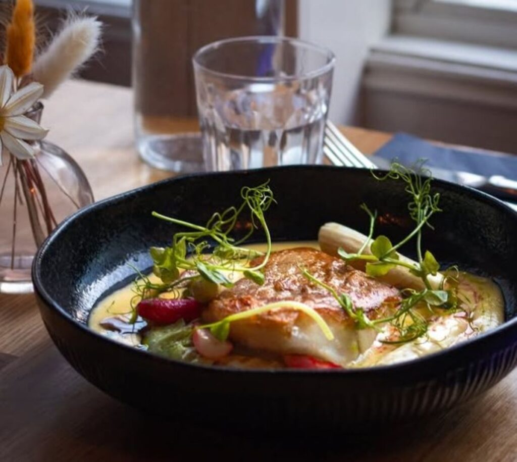 Pan-fried fish with microgreens and vegetables in black skillet, bright natural lighting from restaurant window creating casual atmosphere