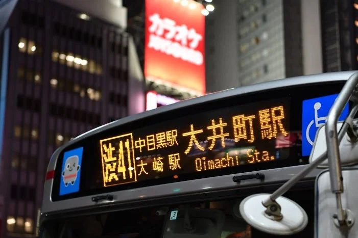 Close-up of an illuminated destination display on a Tokyo bus at night, showing Japanese text and station names in English. Neon signs visible in the background.