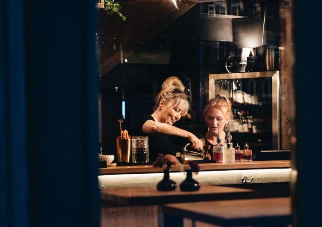 Two blonde bartenders preparing cocktails behind illuminated bar counter in dimly lit restaurant, creating warm evening atmosphere