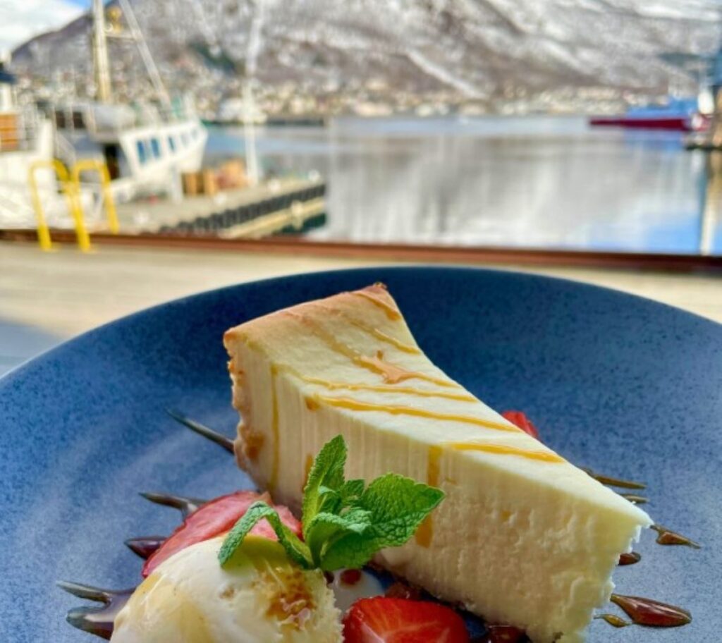Slice of cheesecake with fresh strawberries and mint on blue plate, fishing boats visible through restaurant window in Tromsø harbour