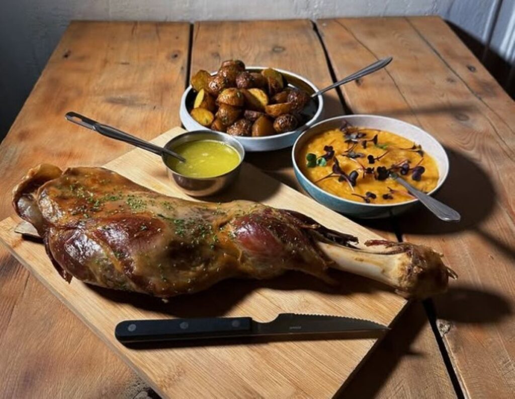 Whole roasted duck leg on wooden board with roasted potatoes, green sauce and orange carrots, rustic Nordic dining presentation
