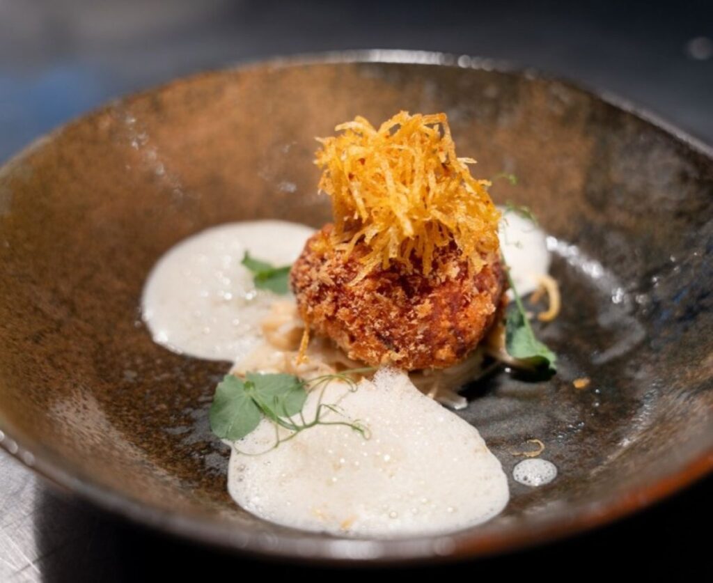 Crispy croquette with golden potato nest garnish and white foam sauce on dark ceramic bowl, showcasing fine dining presentation