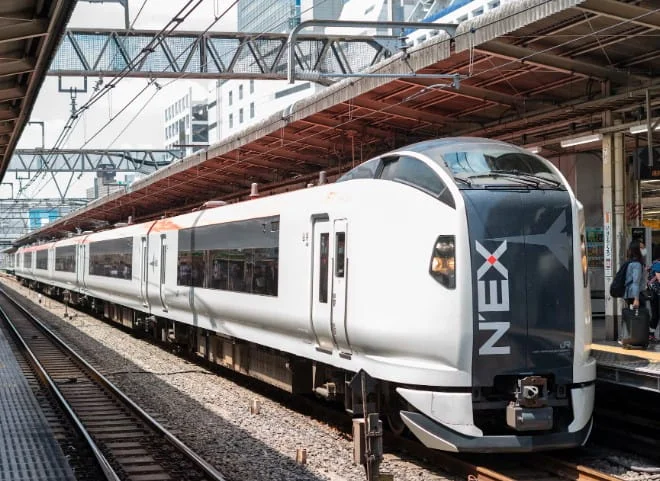A sleek white NEX express train stopped at a Japanese railway platform. The train has a streamlined nose and "NEX" branding visible on its side.