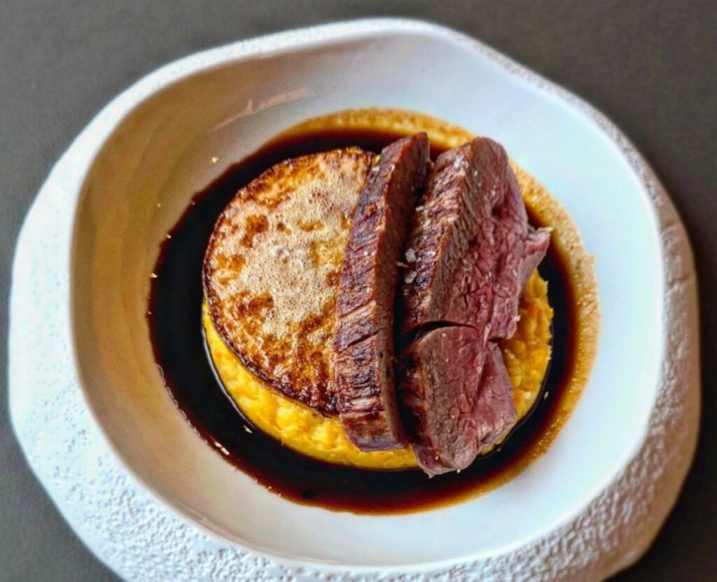 Seared duck breast sliced medium-rare with golden potato fondant and rich red wine jus in white ceramic bowl