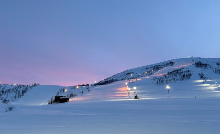 Evening view of illuminated ski slopes at Tromsø Alpinpark, Norway. Snow-covered hills with lit ski runs and lift lines contrast against a pastel purple and blue twilight sky, creating a serene winter landscape.