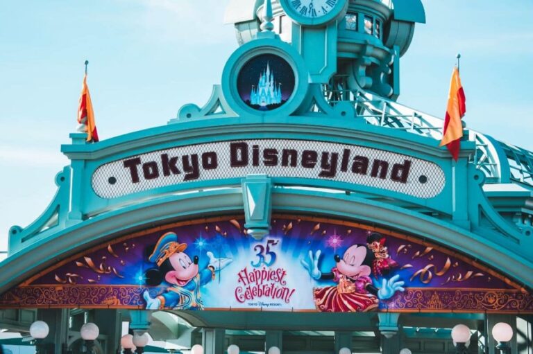 The entrance arch to Tokyo Disneyland featuring turquoise Art Deco-style architecture. The park's name is displayed in black letters on a white background. Above it is a circular medallion containing an illuminated silhouette of Cinderella Castle. Below is a celebratory banner for the park's 35th "Happiest Celebration!" featuring Mickey and Minnie Mouse in festive costumes against a magical blue background with golden confetti. Orange flags flutter at the top corners of the structure, and white paper lanterns decorate the base.
