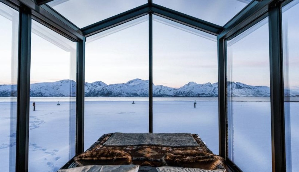 View from inside a glass cabin with a bed under fur throws, looking out through glass walls and roof to a snow covered shore and mountains beyond