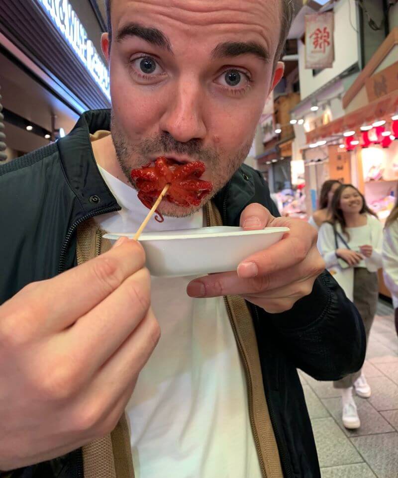 A lively moment at Kyoto’s Nishiki Market, savouring a vibrant skewered octopus, capturing the essence of Japan’s street food culture.