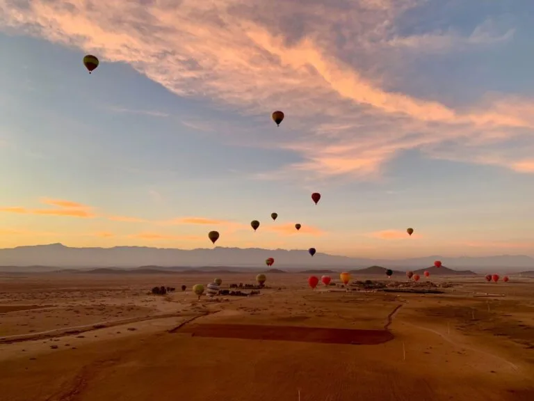 A spectacular sunrise over the Marrakech desert landscape, with dozens of hot air balloons floating at various heights against a pink and orange sky. The desert floor below shows a patchwork of sandy terrain and agricultural plots, whilst the Atlas Mountains rise hazily in the background. The balloons appear in various colours, including red, orange and yellow, creating a dreamy, otherworldly scene