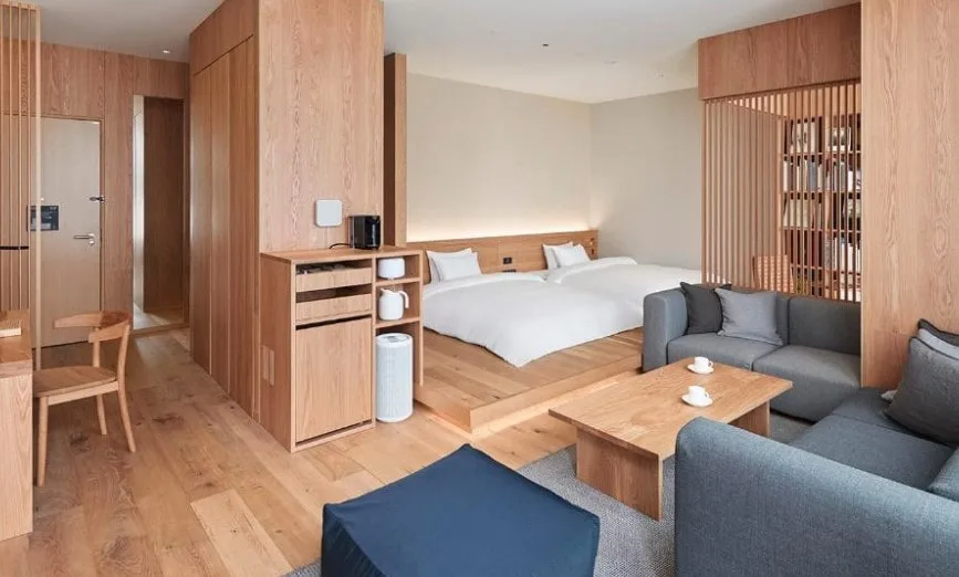 Modern Japanese-inspired room with light oak flooring and panelling. Features twin beds on a raised platform, grey corner sofa, and clever storage solutions. Clean lines and natural materials throughout.