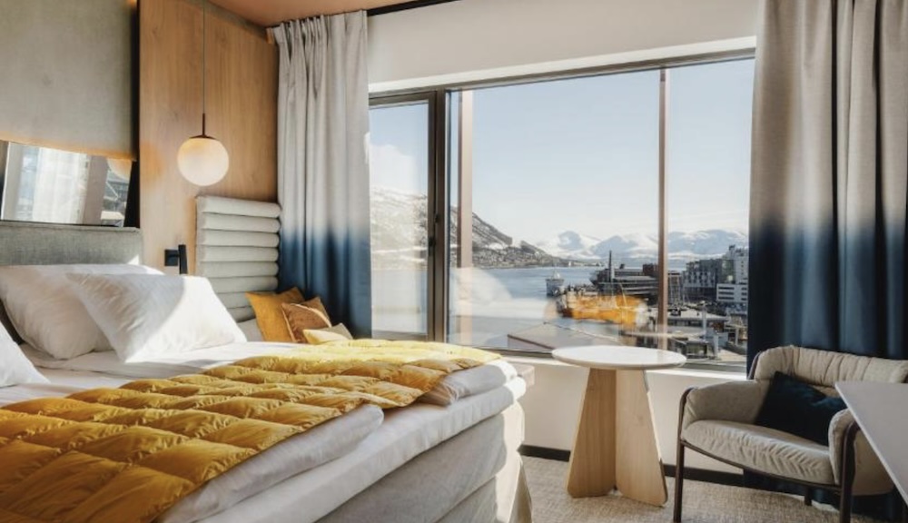 Hotel room with a yellow quilted throw across a large bed, a globe pendant light and floor to ceiling windows looking over a harbour to snowy peaks