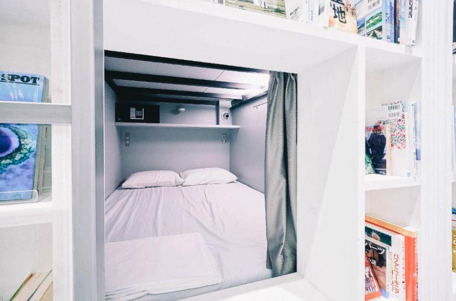 Capsule-style bed nook with white bedding, surrounded by built-in bookshelves filled with manga and travel guides.