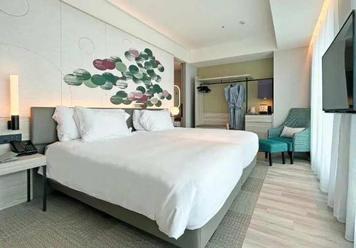 Modern bedroom with abstract leaf wall art, white bedding and turquoise armchairs. Dressing area visible through doorway.