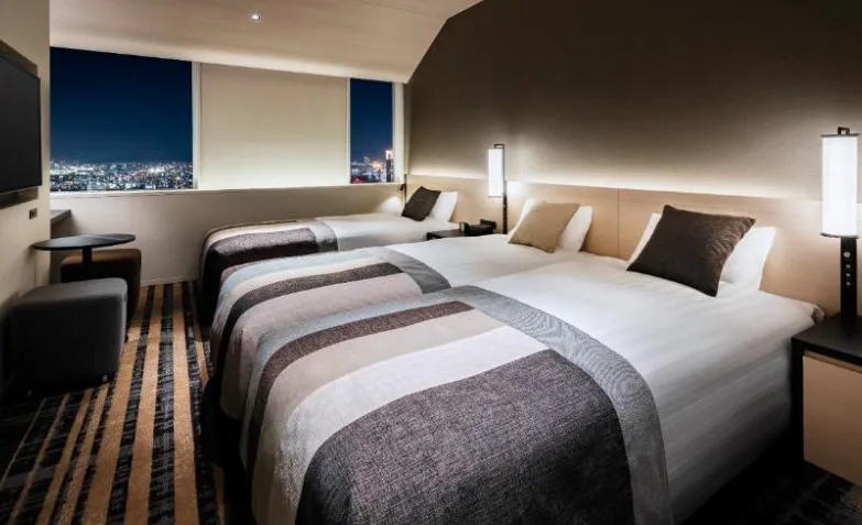 Twin beds with striped grey coverlets in modern room. Night cityscape visible through angular windows. Geometric carpet and wall lighting.