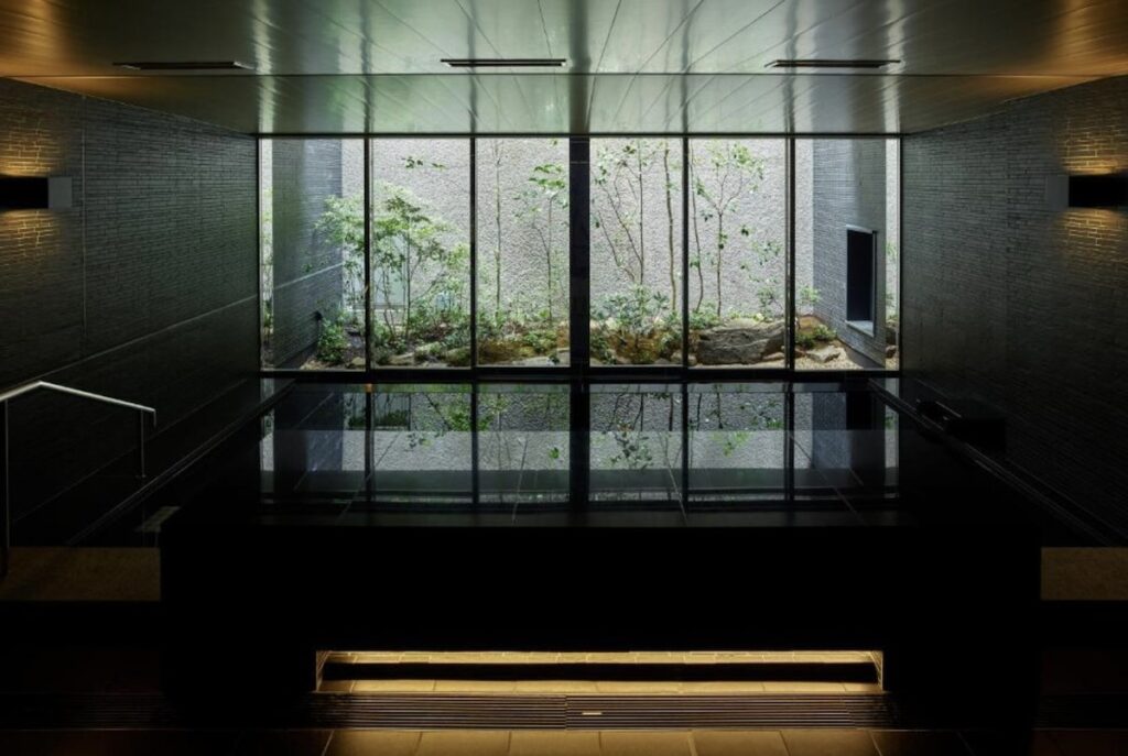 Dark stone onsen bath at Hotel The Celestine Kyoto Gion, with floor-to-ceiling windows overlooking a small illuminated garden of trees and rocks.