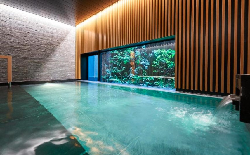 Indoor pool with wooden slatted wall and stone texture panels. Large window overlooks lit bamboo garden.