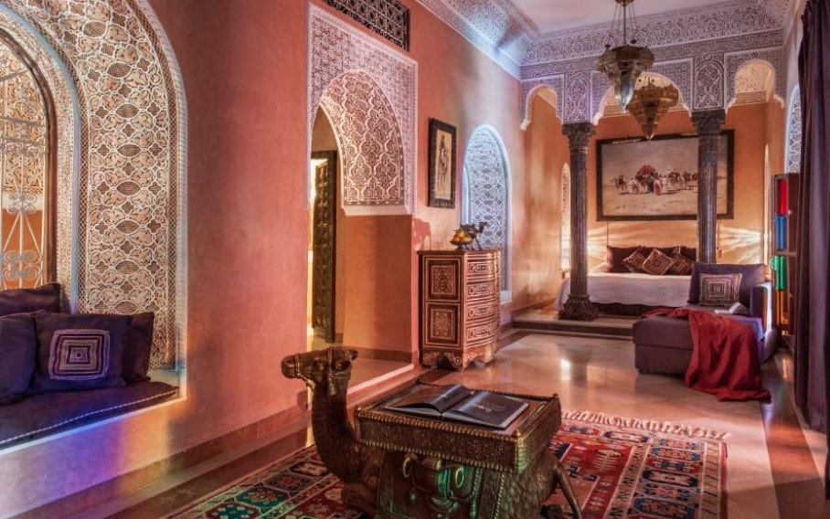 Riad bedroom suite with ornate carved arches, terracotta walls, decorative camel trunk and traditional Moroccan carpets