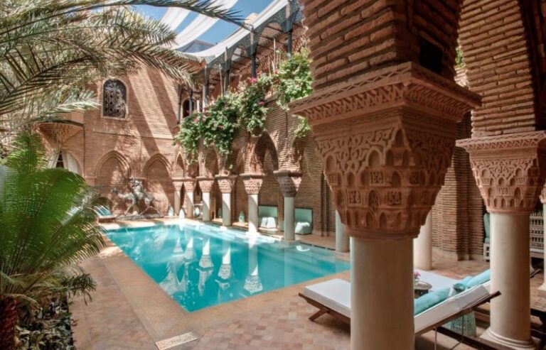 Riad courtyard pool surrounded by brick arches with carved capitals, sunloungers and climbing plants