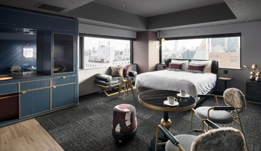 Corner hotel room with city views through large windows. Navy built-in storage, double bed, grey carpet and modern seating with brass accents.