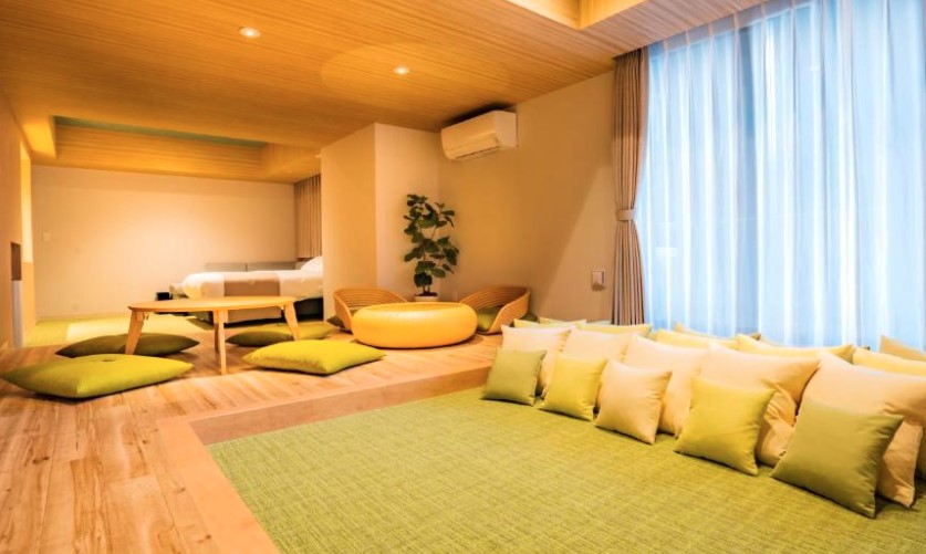 Bright room with wooden floors and raised platform bed. Large green cushions, yellow pouf and cream curtains create casual seating area.