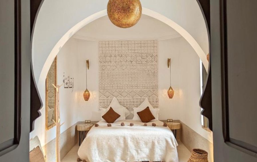 Minimalist riad bedroom through Moorish arch, featuring white linens, bronze pendant lights and geometric wall art
