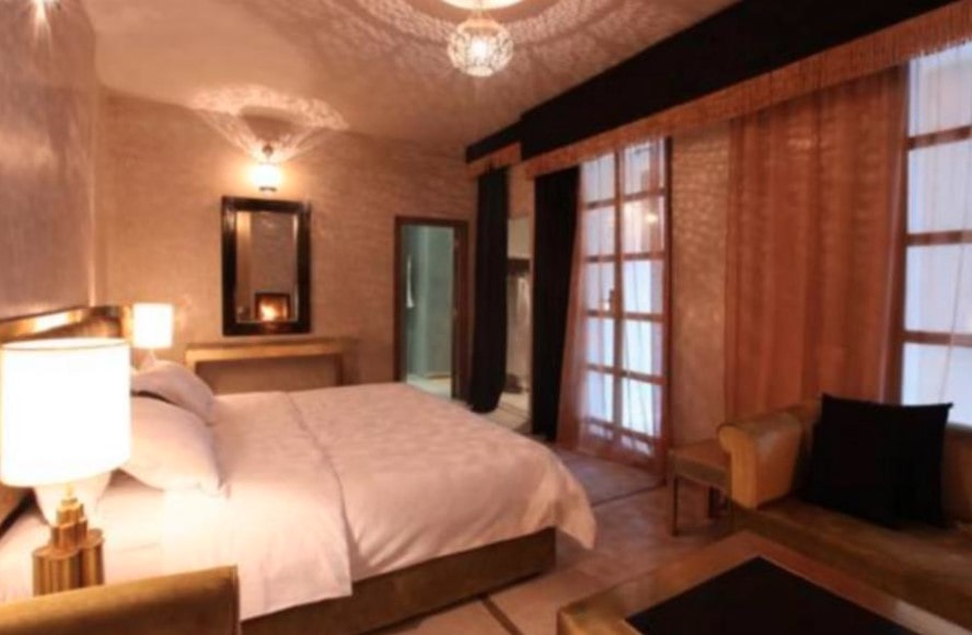 Riad bedroom with white bedding, exposed wooden beams and French doors, lit by soft wall sconces and chandelier