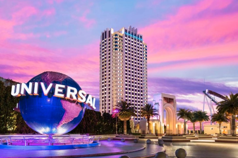 Hotel tower beside illuminated Universal globe at sunset. Pink and purple sky with palm trees lining entrance plaza.