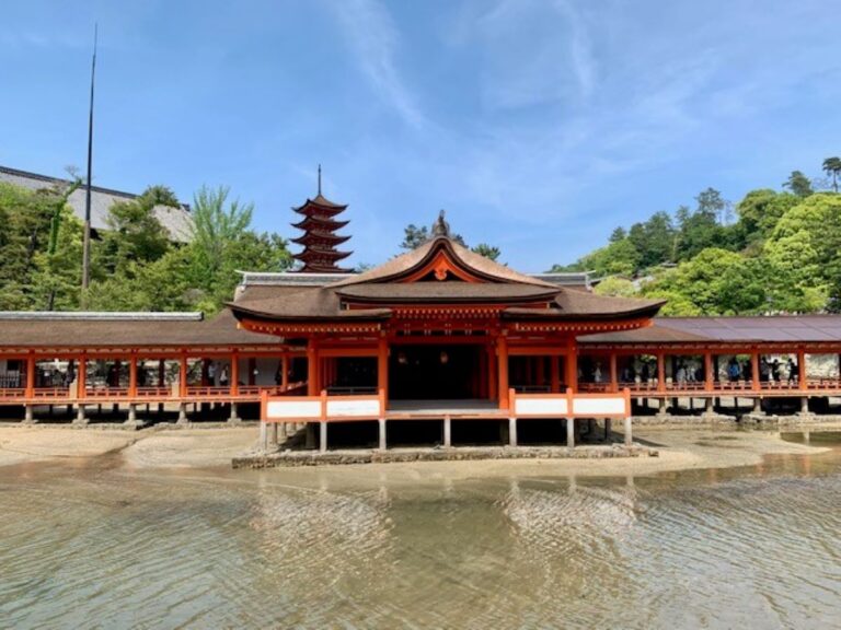 The vermillion-coloured Itsukushima Shrine with its distinctive five-storey pagoda, built over water on stilts at Miyajima, Japan, set against lush greenery and blue skies
