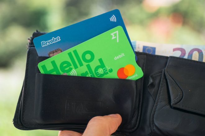 Two payment cards, one blue Revolut and one green Hello card, partially visible in a black leather wallet alongside Euro banknotes