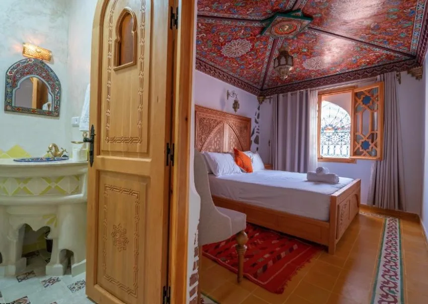 Ornate Moroccan-style bedroom with intricate ceiling, carved wooden door, colourful textiles, and arched architectural details.