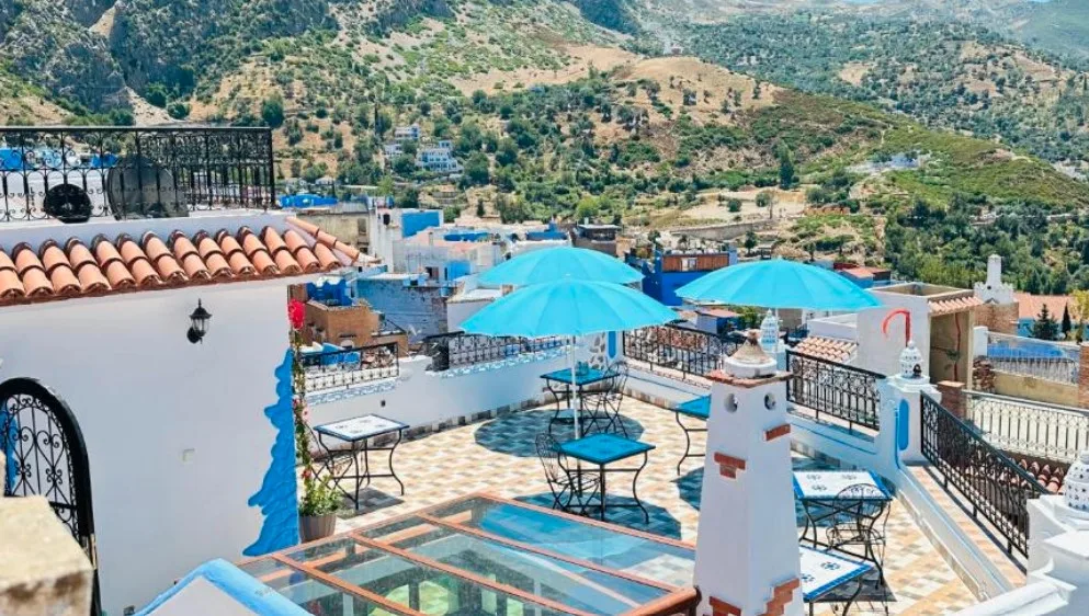 Rooftop terrace with blue umbrellas, mosaic tiles, and mountain village backdrop, showing white buildings and terracotta roofs.