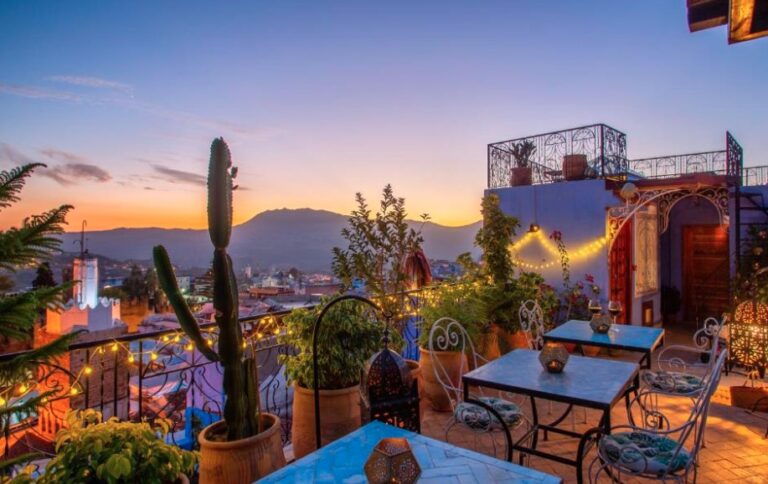 Rooftop terrace at sunset with cacti, fairy lights, wrought-iron furniture, overlooking a mountain town with warm evening sky.