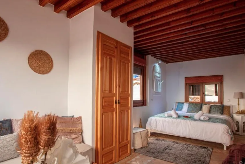 Rustic bedroom with wooden beam ceiling, warm wooden wardrobe, white walls, woven wall decor, and a comfortable bed with cushions.