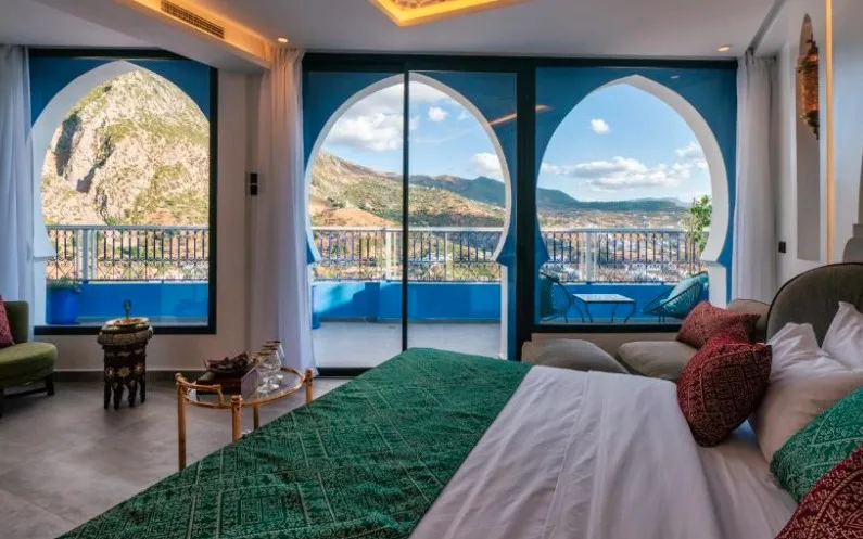 Luxurious bedroom with arched windows overlooking mountainous landscape, green bedspread, and blue balcony seating areas.