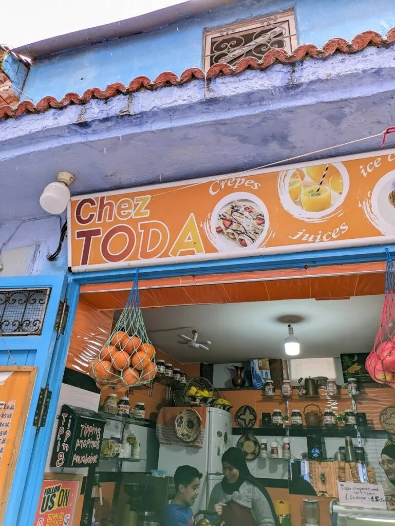 Bright orange shopfront of Chez Toda cafe, with hanging nets of oranges and handwritten signs in Chefchaouen.