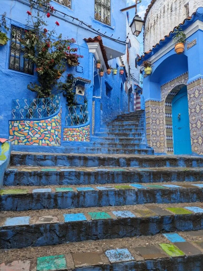 Steep blue-painted steps with colourful mosaic tiles, surrounded by blue-washed walls, intricate metalwork, and flowering plants in Chefchaouen.