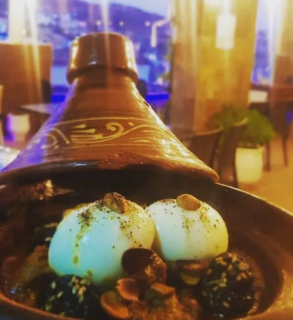 Traditional ceramic tagine with two poached eggs, spices, and herbs, set in a softly lit restaurant interior.