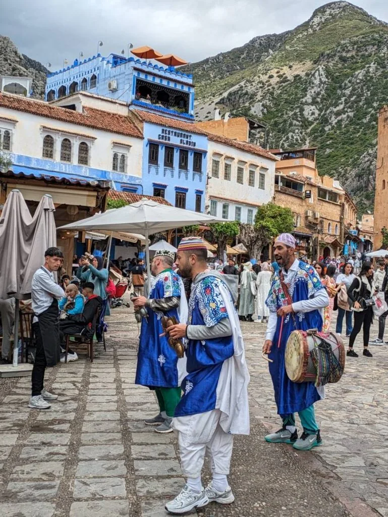 Traditional musicians in blue and white attire performing in a cobblestone street, with mountain backdrop.