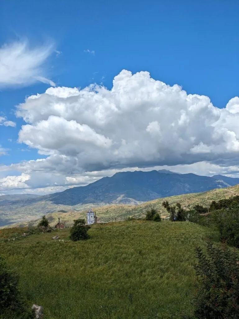 Grassy meadow with a white tower, mountains and dramatic cloud formations in the distance.