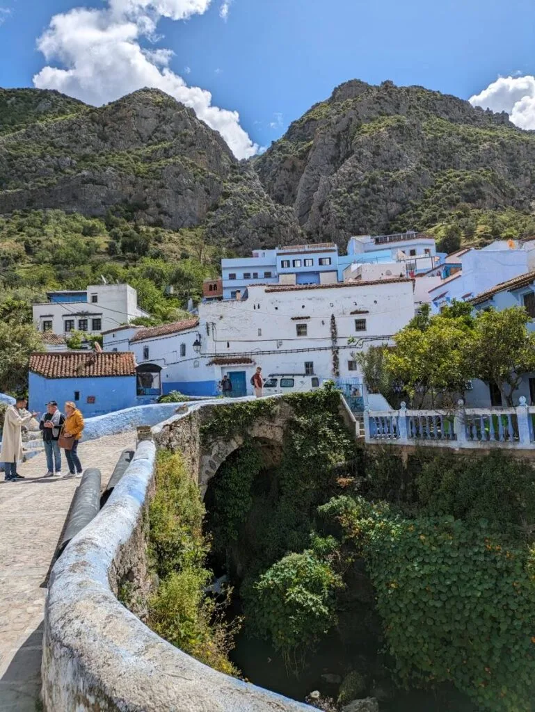 Stone bridge over a green ravine with blue-and-white buildings and rocky mountains in Chefchaouen.