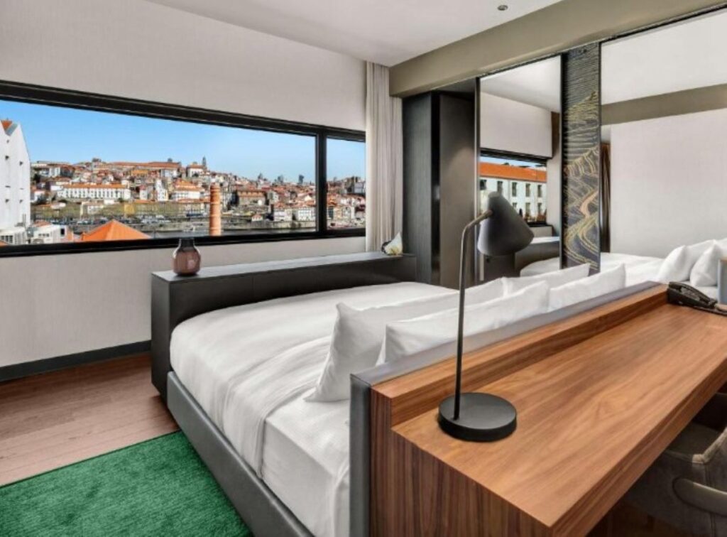 Contemporary hotel room with panoramic window overlooking Porto's riverside district, featuring wooden desk and green accent rug